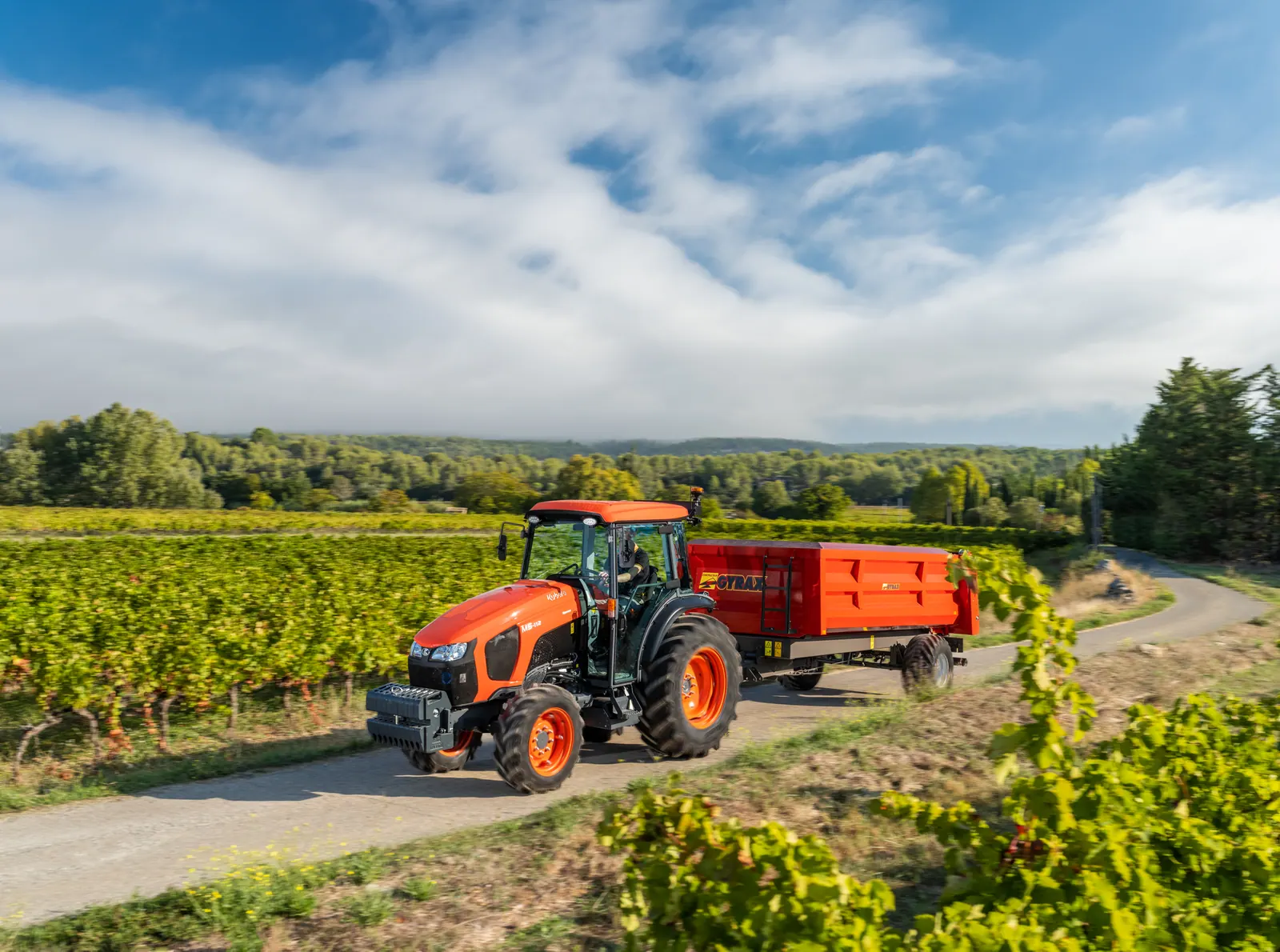Uski traktor Kubota M5-112 N s kabinom vozi prikolicu po putu uz vinograd