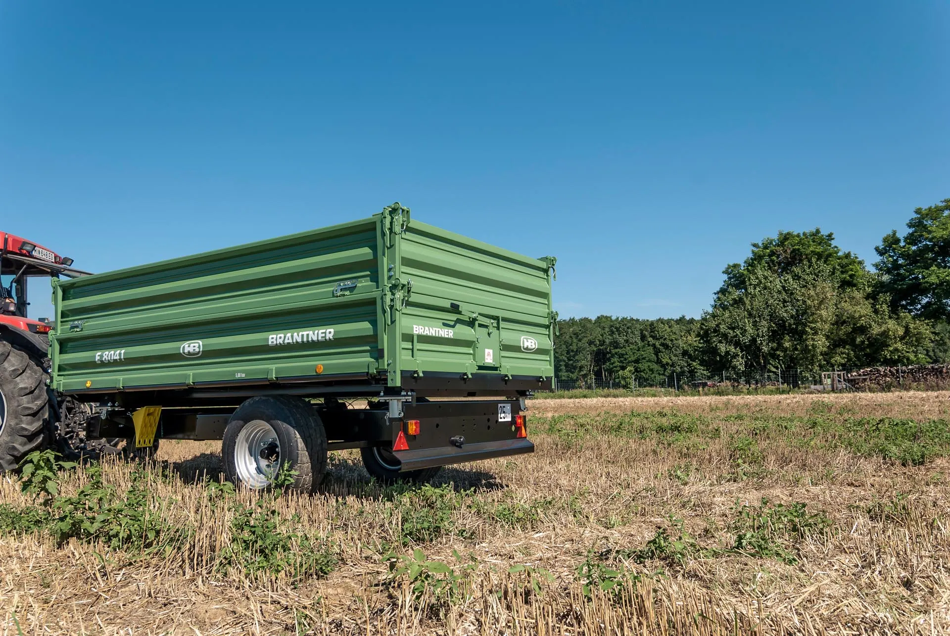 Brantner E 8041 jednoosovinska prikolica prikopčana na crveni traktor, na strništu uz drveće