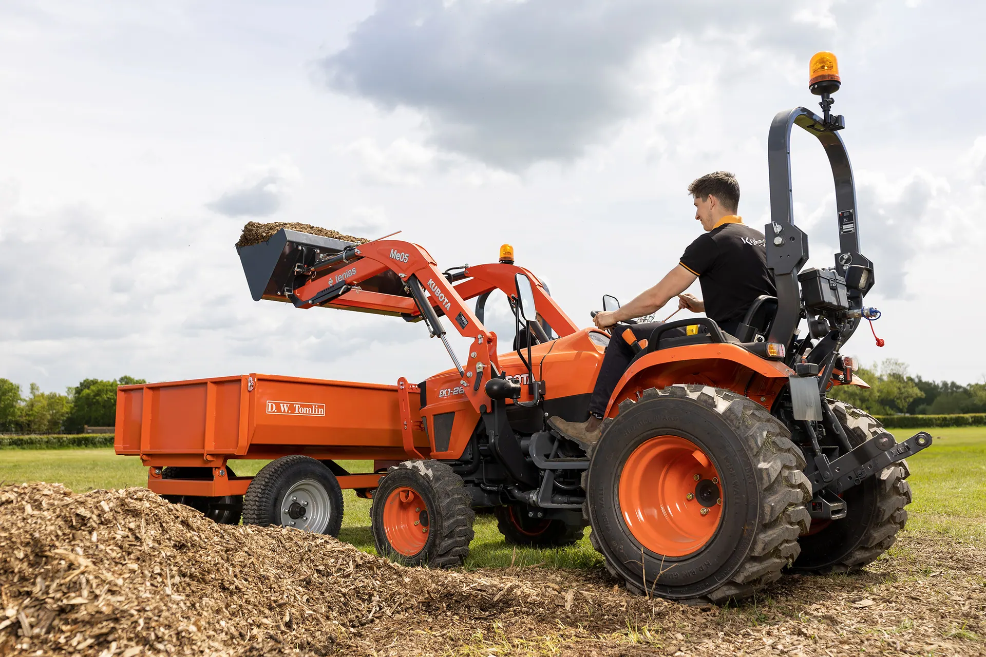 Kubota EK1 s prednjim utovarivačem istresa sječku u narančastu prikolicu
