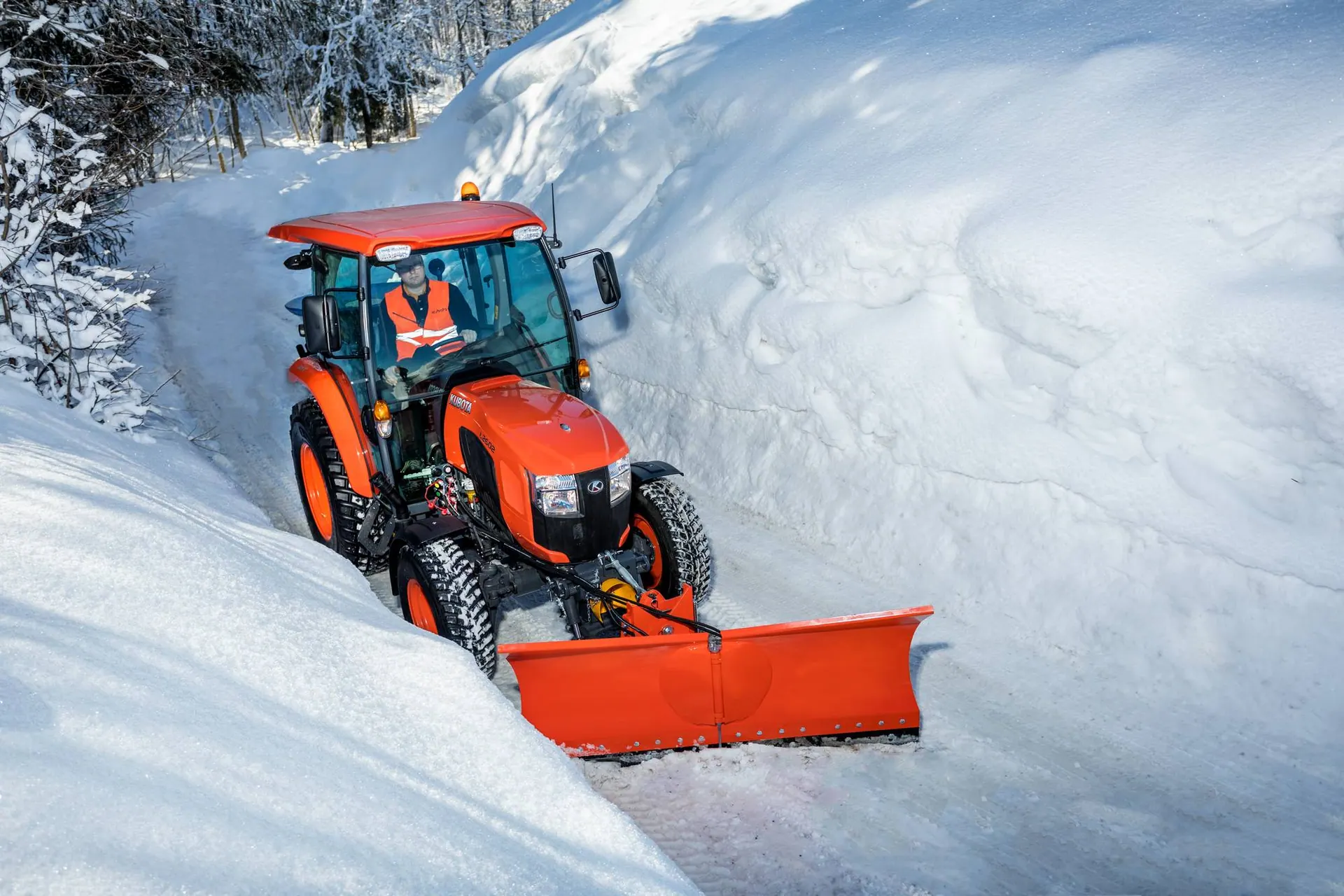 Kubota L2 s kabinom i prednjom ralicom čisti snijeg na šumskoj cesti između snježnih nasipa