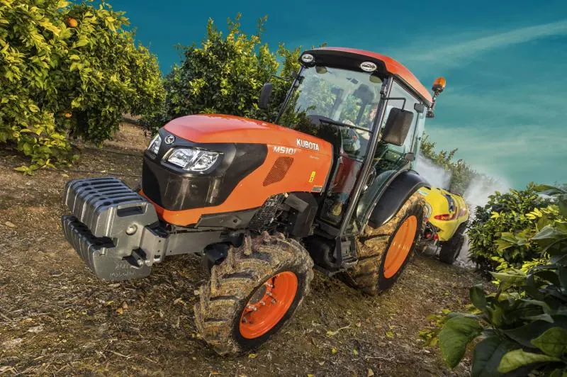 Uski traktor Kubota M5002 Narrow s kabinom i prednjim utezima u nasadu naranči, iza prskalica