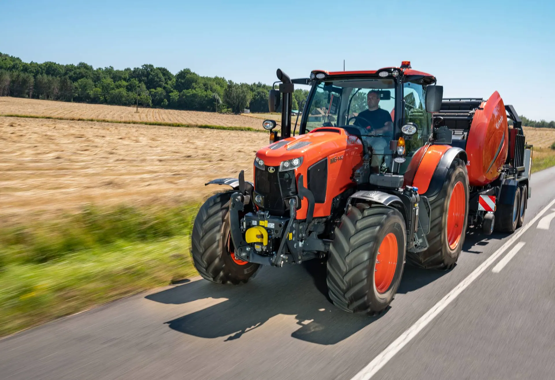 Kubota M6 s kabinom vuče prešu za okrugle bale cestom uz pokošeno polje