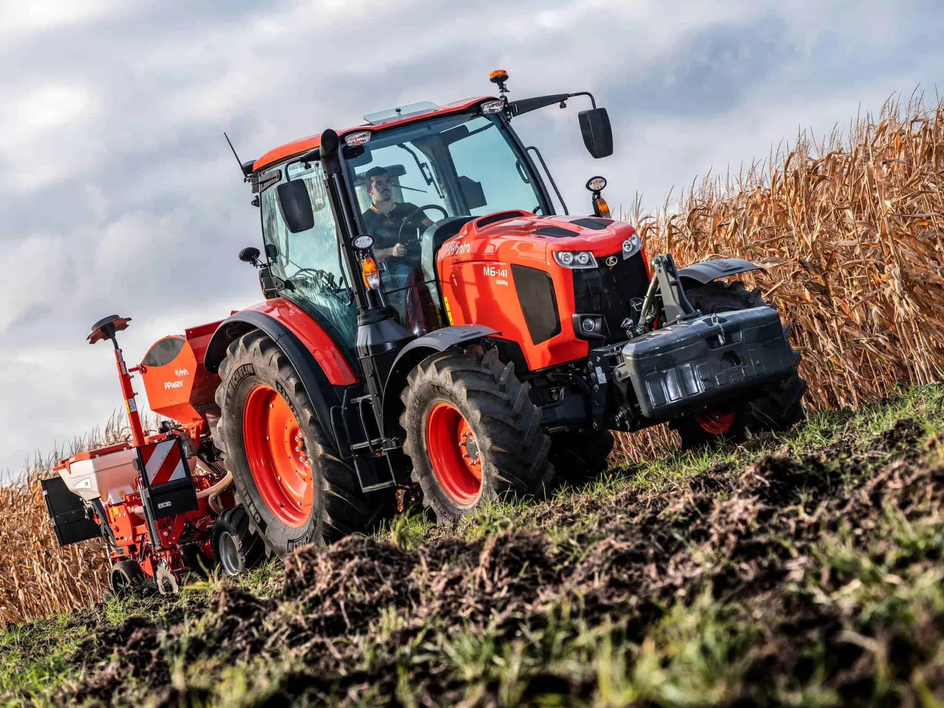 Kubota M6 Utility s prednjim utegom i sijačicom Kubota vozi po polju uz kukuruzište, pogled odozdo